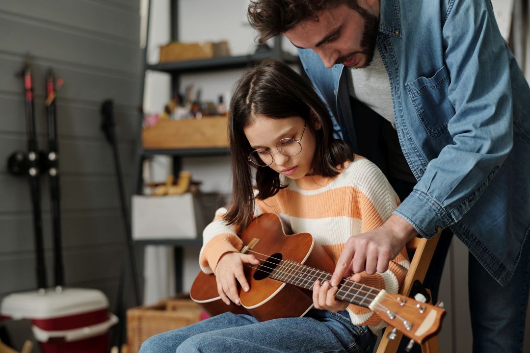 Little girl in eyeglasses learning to play guitar while young man bending over her and keeping finger on one of strings during Guitar Class