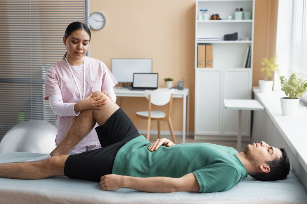 A physical therapist examines a man's bent knee as he lies on a treatment table in a rehabilitation-focused chiropractic care clinic room.