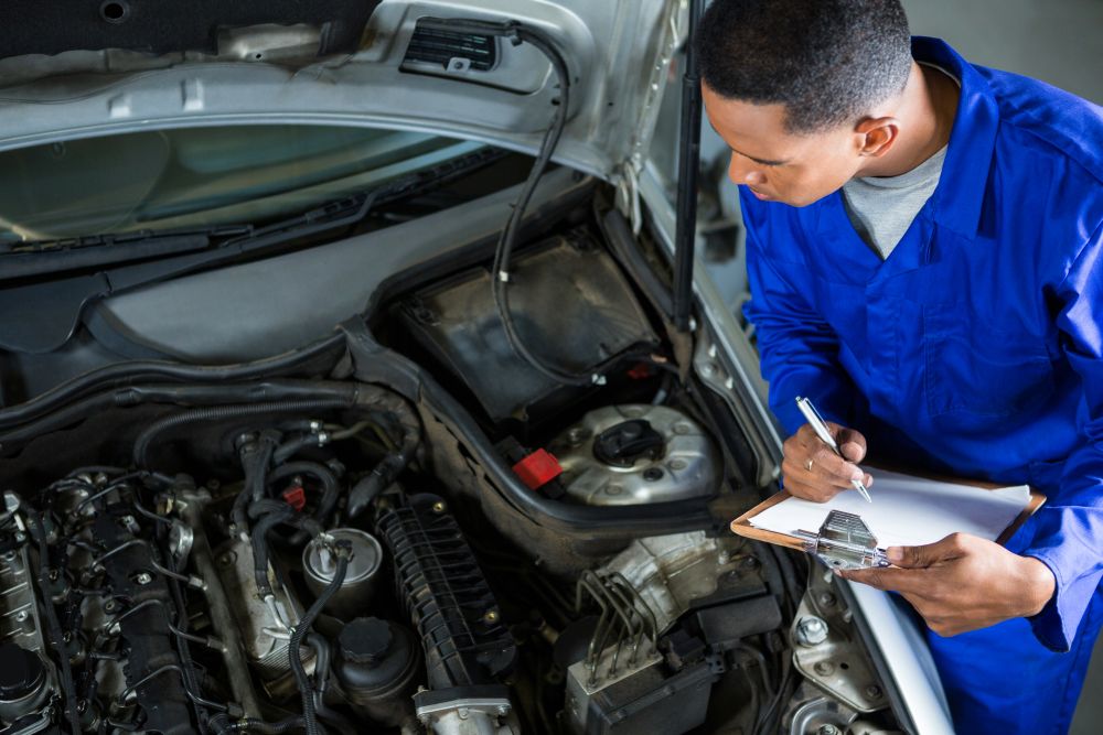 Mechanic in blue coveralls examines a car engine and takes notes on a clipboard while standing beside the open hood, demonstrating exactly what to know before getting an engine overhaul.